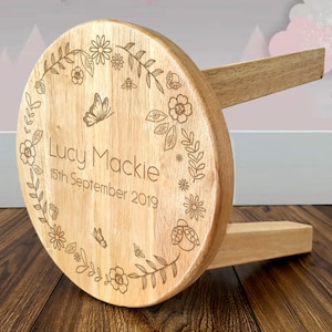 May include: A wooden stool with a circular seat, featuring engraved floral and butterfly motifs around the edge. The name "Lucy Mackie" and the date "15th September 2019" are inscribed. The stool has two wooden legs.