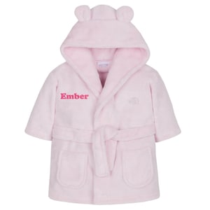 May include: A light pink hooded baby bathrobe with bear ears on the hood. The robe has a matching belt, two front pockets, and the name "Ember" embroidered in pink. A small logo is embroidered on the right side.