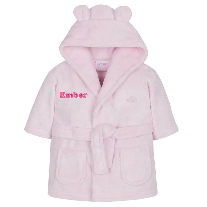 May include: A light pink hooded baby bathrobe with bear ears on the hood. The robe has a matching belt, two front pockets, and the name "Ember" embroidered in pink. A small logo is embroidered on the right side.
