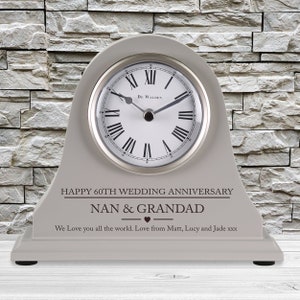 May include: Gray wooden mantel clock with a white face and black Roman numeral hour markers. The clock has a personalized inscription that reads "Happy 60th Wedding Anniversary Nan & Grandad We Love you all the world. Love from Matt, Lucy and Jade xxx".