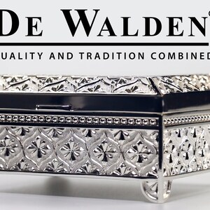 May include: A silver metal trinket box with an ornate design and a hinged lid. The box is sitting on a white surface. The text "DE WALDEN" and "QUALITY AND TRADITION COMBINED" is printed on the white surface above the box.