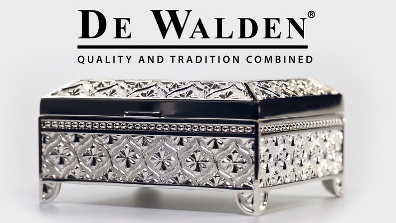 May include: A silver metal trinket box with an ornate design and a hinged lid. The box is sitting on a white surface. The text "DE WALDEN" and "QUALITY AND TRADITION COMBINED" is printed on the white surface above the box.