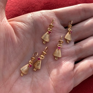 May include: Five gold charms in the shape of a girl holding a red heart. The charms are on a hand.