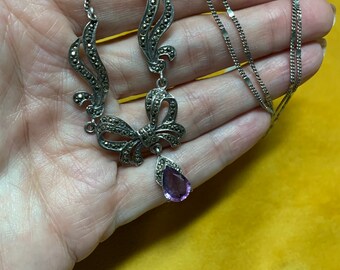Può includere: Una collana color argento con un design a fiocco e volute, impreziosita da pietre di marcasite. Una gemma viola a forma di goccia pende dall'arco. La collana è esposta su una superficie gialla.