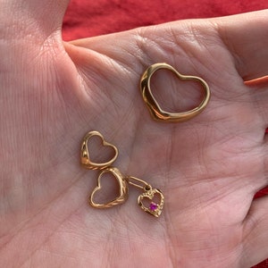 May include: Three gold heart-shaped pendants. Two of the pendants are open and one has a small pink gemstone in the center.
