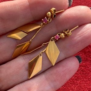 May include: A pair of gold earrings with a geometric design. Each earring features three gold diamond shapes with a small pink gemstone at the top.