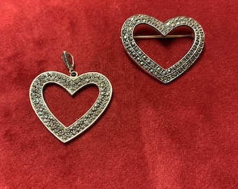 Op de afbeelding: Twee zilveren hartvormige hangers met een pavé-zetting van kleine, donkergrijze stenen. De hanger aan de linkerkant is een bedel met een lus aan de bovenkant. De hanger aan de rechterkant is een broche met een pin aan de achterkant.