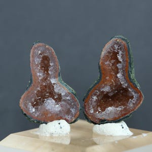 May include: Two matching, polished, geode halves. The geodes have a dark green outer layer, a reddish-brown interior, and a sparkling crystal center. They are mounted on small, white, irregular bases, and sit on a light-colored surface.