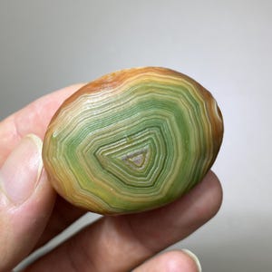 May include: A polished, oval-shaped agate stone featuring concentric bands of colour. The centre displays shades of light green and white, transitioning outwards into layers of yellow, orange, and brown. The surface is smooth with a glossy finish.
