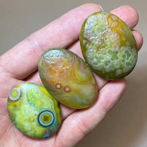 May include: Three polished oval gemstones in shades of green, yellow, and orange. The stones have unique patterns, including circular and swirling designs. The gemstones are held in a hand, showcasing their natural beauty and variations in colour and texture.