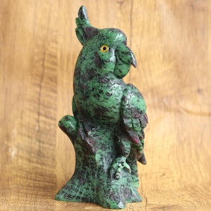 Könnte beinhalten: Eine geschnitzte Papagei-Skulptur aus Stein in Grün- und Burgundertönen, die auf einem Baumstamm sitzt. Der Papagei hat ein detailliertes Design mit gelben Augen und einem gebogenen Schnabel. Die Skulptur ist ein dekorativer Gegenstand.