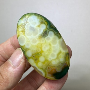 Puede incluir: Una piedra preciosa pulida de forma ovalada con una superficie translúcida que presenta patrones en capas de orbes circulares blancos, amarillos y verde oscuro. La piedra muestra una textura suave y brillante con bandas concéntricas.