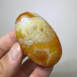 May include: An oval-shaped polished agate stone featuring a vibrant colour palette of golden yellow, deep orange, and translucent white. The surface displays intricate, concentric circular banding patterns and organic, layered formations throughout.