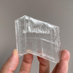 以下が含まれることがあります： 長方形の形をした透明なセレン石の結晶。 半透明の石には、目に見える縞模様とわずかに粗い質感があります。 結晶は、ニュートラルな背景に対して手で保持されています。