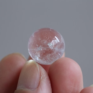 May include: A small, spherical, translucent clear quartz crystal ball with visible internal inclusions and natural fractures. The stone has a smooth, polished surface that reflects light, showcasing a pale, icy appearance with subtle cloudy white patterns.