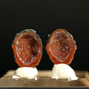 May include: Two halves of a geode, showcasing a vibrant interior of sparkling crystals. The outer edges are a deep orange-red, with a dark, almost black, border. Each half is mounted on a white, textured base, set against a black backdrop.