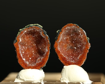 May include: Two halves of a geode, showcasing a vibrant interior of sparkling crystals. The outer edges are a deep orange-red, with a dark, almost black, border. Each half is mounted on a white, textured base, set against a black backdrop.