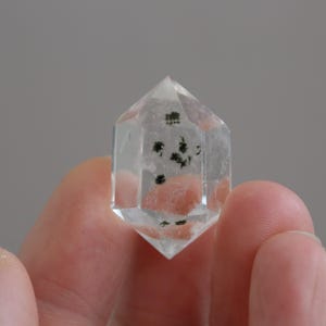 Könnte beinhalten: Ein klarer Quarz-Kristall mit dunkelgrünen Einschlüssen. Der Kristall hat eine sechseckige Form mit einer spitzen Spitze und wird zwischen den Fingern gehalten. Die Einschlüsse erscheinen als kleine, dunkle Cluster im transparenten Quarz.