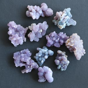 May include: A collection of 11 purple and grey agate stones with a grape-like texture. The stones are arranged on a dark grey surface.