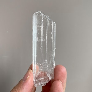 Può includere: Un cristallo chiaro e allungato con una superficie a coste. Il cristallo è tenuto in mano, mostrando la sua qualità traslucida e i bordi naturali e irregolari. Il colore del cristallo è una tonalità pallida, quasi incolore e trasparente.