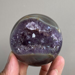 Könnte beinhalten: Eine polierte Amethyst-Geodenkugel. Das Äußere ist ein durchscheinendes Braun und Grau, das ein tiefviolettes Inneres mit funkelnden Amethystkristallen freilegt. Die Geode wird in einer Hand gehalten.