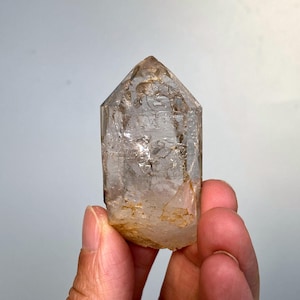 Peut inclure: Une pointe de cristal de quartz clair avec une base naturelle et rugueuse. Le cristal est tenu dans une main, mettant en valeur ses teintes transparentes et brun clair. Les facettes du cristal captent la lumière.