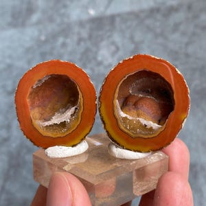 Könnte beinhalten: Zwei Hälften einer kreisförmigen Achat-Geode mit leuchtenden konzentrischen Ringen in Orange, Gelb und Dunkelrot. Das Innere jedes Stücks zeigt einen Hohlraum, der mit strukturierten, goldbraunen Kristallformationen und weißen Mineralablagerungen ausgekleidet ist.