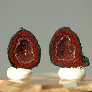 May include: Two halves of a geode, showcasing a deep red interior with crystalline formations. The outer layer is dark, with a green and black textured surface. Each half rests on a small, white, rounded base, set against a neutral background.