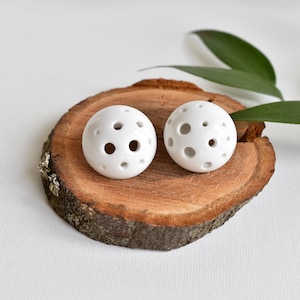 Op de afbeelding: Twee witte keramische oorbellen met een ronde vorm en meerdere ronde uitsparingen. De oorbellen worden gepresenteerd op een houten oppervlak.