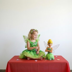 May include: A low-poly paper sculpture of a fairy with yellow hair, a green bodice, and translucent wings, positioned next to a child wearing a green sequined dress with matching fairy wings and a floral headband, both set against a plain white background.