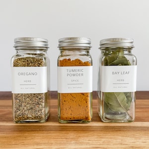 May include: Three glass spice jars with silver lids, each labeled with white labels. The jars contain oregano, turmeric powder, and bay leaves. The labels read "OREGANO HERB ALL NATURAL", "TUMERIC POWDER SPICE ALL NATURAL", and "BAY LEAF HERB ALL NATURAL".