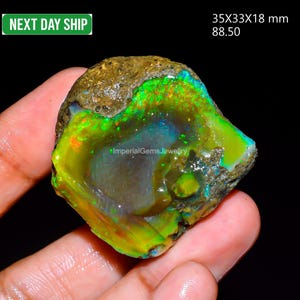 May include: A raw, unpolished opal gemstone featuring vibrant flashes of neon green, yellow, and orange play-of-color against a dark, earthy matrix. The stone measures 35x33x18 mm and displays a natural, irregular shape with a textured surface.