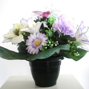 Peut inclure: Un arrangement de fleurs artificielles dans un pot noir, composé de fleurs violettes, blanches et vertes. L'arrangement comprend des lys, des œillets et des marguerites.
