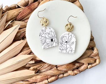 May include: A pair of white, arch-shaped earrings with a black floral design. Each earring has a gold-colored, textured disc at the top and a gold-colored hook. The earrings are displayed on a white, round plate, set on a woven, brown surface.