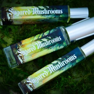 May include: Three rectangular perfume bottles with silver caps are displayed on a bed of green moss. Each bottle has a label with the words "Sugared Mushrooms" in a decorative font. The labels feature a colorful, artistic design.