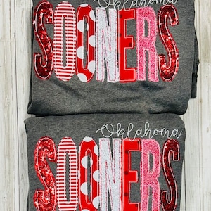 May include: Two gray sweatshirts with the words "Oklahoma Sooners" in red, white, and pink letters with sequins. The letters are decorated with red and white stripes, white polka dots, and pink sequins.