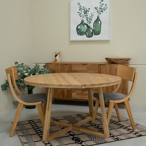 Op de afbeelding: Een ronde houten eettafel met een natuurlijke houten afwerking en een grijs gestoffeerde stoel. De tafel heeft een blad dat kan worden uitgebreid om hem groter te maken. De tafel staat op een tapijt met een zwart-wit geometrisch patroon.
