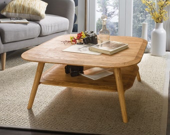May include: A light wood coffee table with a lower shelf. The table has a square top with rounded edges and four legs. The table is sitting on a light beige rug in a living room.