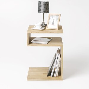 May include: A light wood side table with a unique S-shaped design. The table has two shelves, one for displaying a lamp and a picture frame, and the other for storing books and magazines.