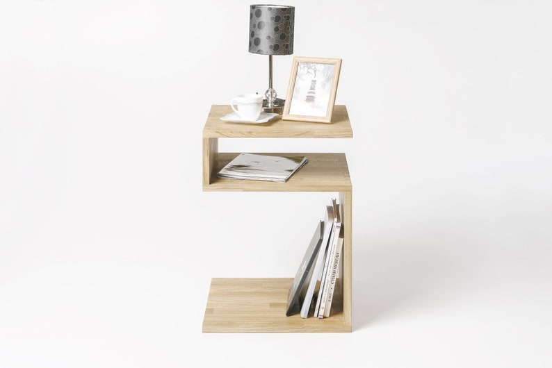 May include: A light wood side table with a unique S-shaped design. The table has two shelves, one for displaying a lamp and a picture frame, and the other for storing books and magazines.