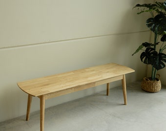 Puede incluir: Un banco de madera clara con un diseño sencillo. El banco tiene una tapa rectangular y cuatro patas. El banco está sentado en un suelo de hormigón.