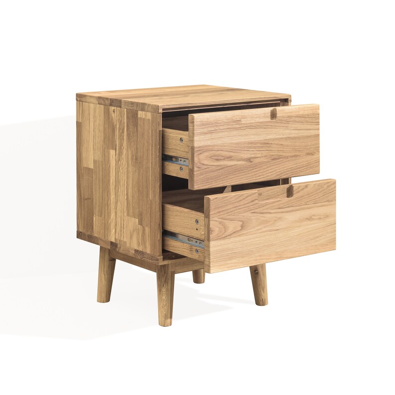 May include: A light-colored wooden nightstand with two drawers, one of which is open. The nightstand has a rectangular shape and sits on four short legs. The wood grain is visible, giving it a natural look.