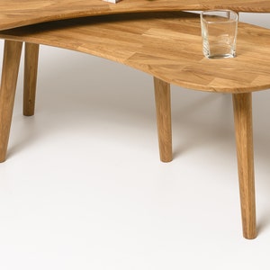 May include: A set of two wooden nesting coffee tables with a unique, organic shape. The tables are made of light brown wood and have a natural finish. The top table is slightly smaller than the bottom table and sits on top of it. The tables are perfect for a modern living room or a contemporary home.