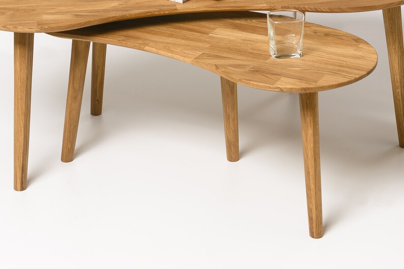 May include: A set of two wooden nesting coffee tables with a unique, organic shape. The tables are made of light brown wood and have a natural finish. The top table is slightly smaller than the bottom table and sits on top of it. The tables are perfect for a modern living room or a contemporary home.