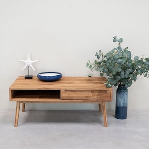 May include: A wooden coffee table with a drawer and a shelf. The table has a light brown finish and is sitting on a gray floor. There is a blue bowl and a white starfish on the table. A vase with a leafy green plant is on the right side of the table.