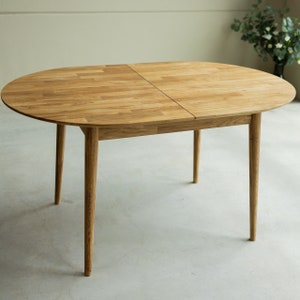 Pu&ograve; includere: Un tavolo da pranzo ovale in legno chiaro con un'estensione a foglia. Il tavolo ha quattro gambe sottili e una finitura in legno naturale.