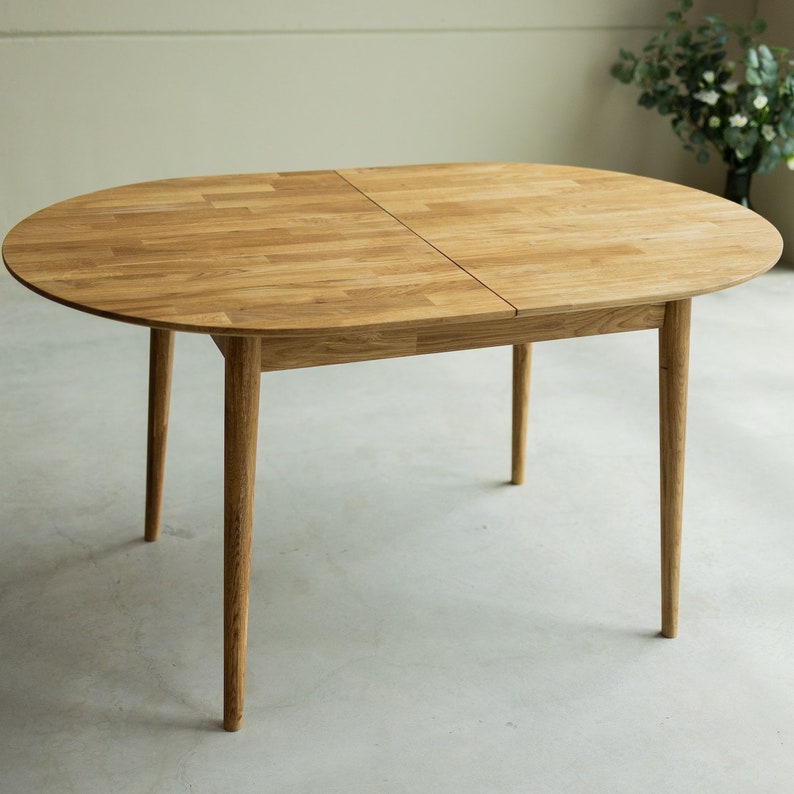 Pu&ograve; includere: Un tavolo da pranzo ovale in legno chiaro con un'estensione a foglia. Il tavolo ha quattro gambe sottili e una finitura in legno naturale.