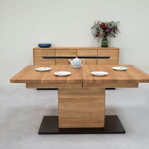 Op de afbeelding: Een eettafel van licht hout met een zwarte basis en vier witte borden en een witte theepot op het oppervlak. De tafel is gedekt voor een maaltijd.