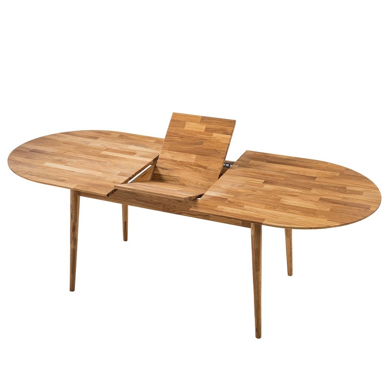 Pu&ograve; includere: Un tavolo da pranzo ovale in legno di colore marrone chiaro con un'estensione a foglia. Il tavolo ha una finitura in legno naturale e quattro gambe affusolate.