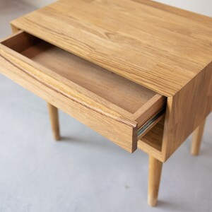 May include: A light wood nightstand with a single drawer that is open. The drawer has a simple design and is made of the same wood as the nightstand. The nightstand has four legs.